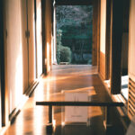 Sunlight casts warm shadows in the wooden hallway of a traditional Japanese house, leading to a garden view at the end.
