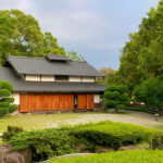 Traditional Japanese house with a tiled roof and wooden sliding doors, surrounded by manicured greenery and trees.
