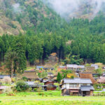 Traditional Japanese mountain village with thatched-roof houses nestled at the edge of a dense forest.