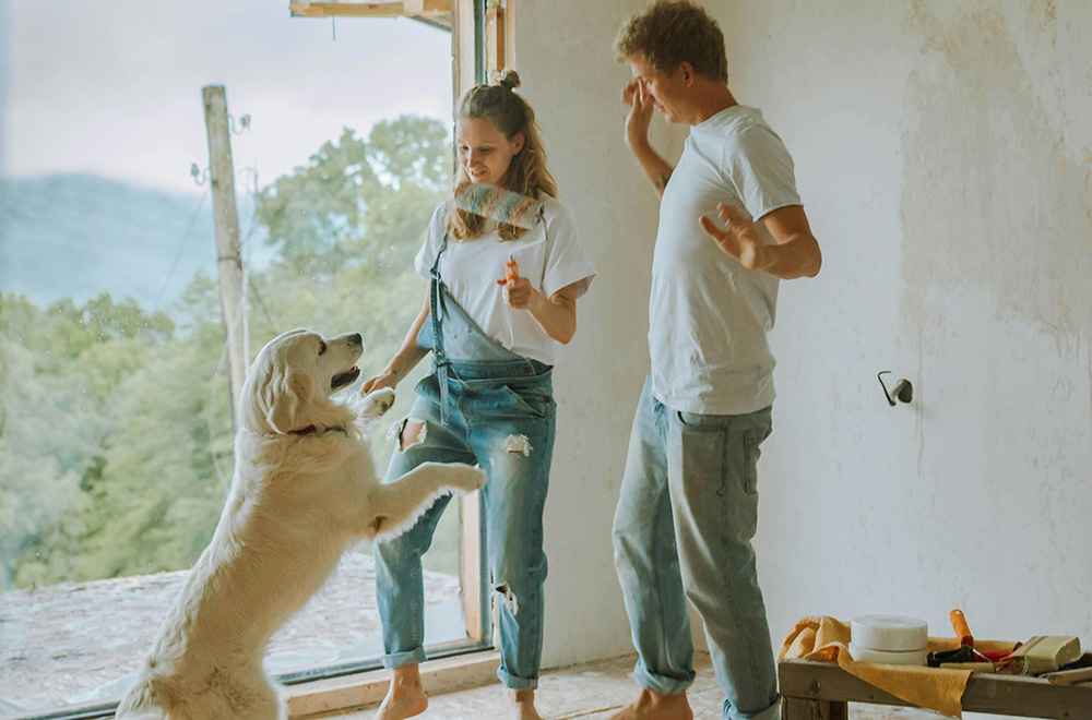 A couple dances with a golden retriever in a partially renovated room with an open doorway overlooking greenery