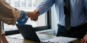 Two people are shaking hands over a desk with a laptop, clipboard, and documents in an office setting