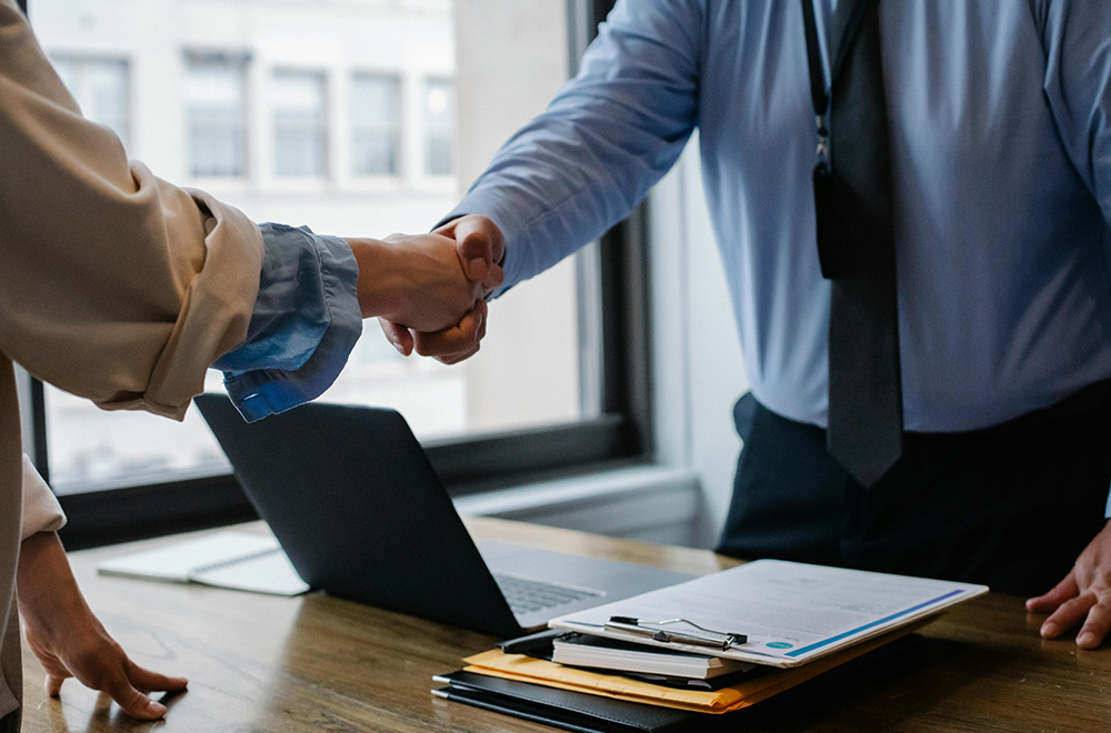 Two people are shaking hands over a desk with a laptop, clipboard, and documents in an office setting