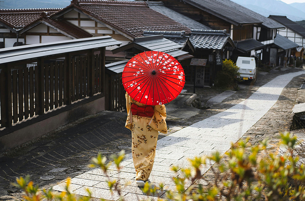 A woman strolls along a path, holding a vibrant red umbrella against the backdrop of a serene landscape