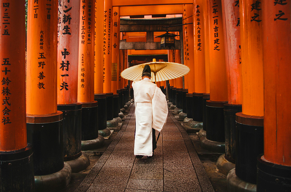 A man in a white robe strolls along a walkway adorned with vibrant orange pillars, creating a serene atmosphere