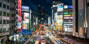 Night view of a busy street in Shinjuku, Tokyo with light trails from traffic and illuminated skyscrapers.