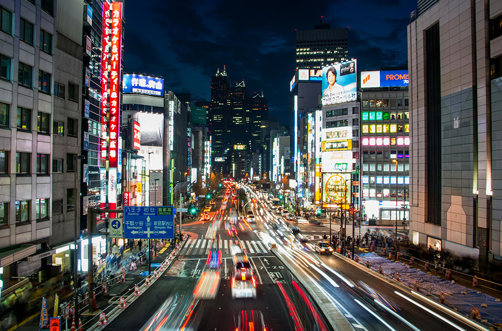 A bustling Tokyo street at night, illuminated by vibrant lights and filled with traffic, showcasing the city's lively atmosphere
