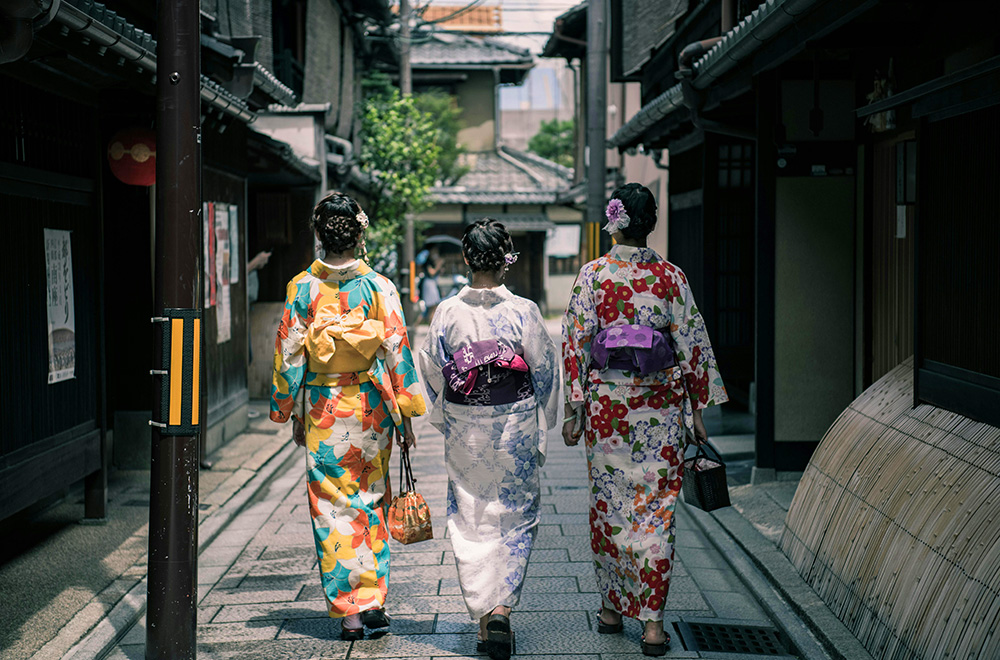 Three women in colorful kimonos stroll through a narrow alley lined with traditional Japanese houses