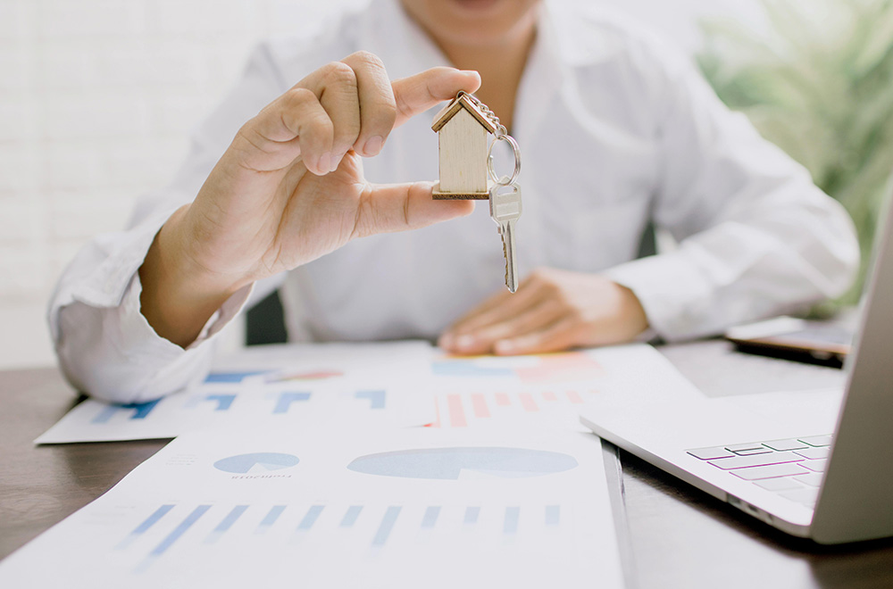 A woman proudly holds a house key in front of a model home, symbolizing her journey in real estate.