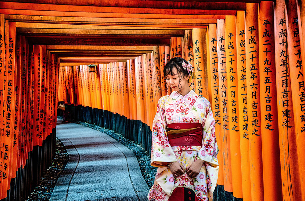 A young woman in a floral kimono stands gracefully among the vibrant red torii gates of Fushimi Inari Shrine in Kyoto, Japan.