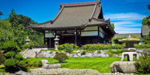 A traditional Japanese temple surrounded by a manicured garden with stone lanterns and lush greenery under a clear blue sky.