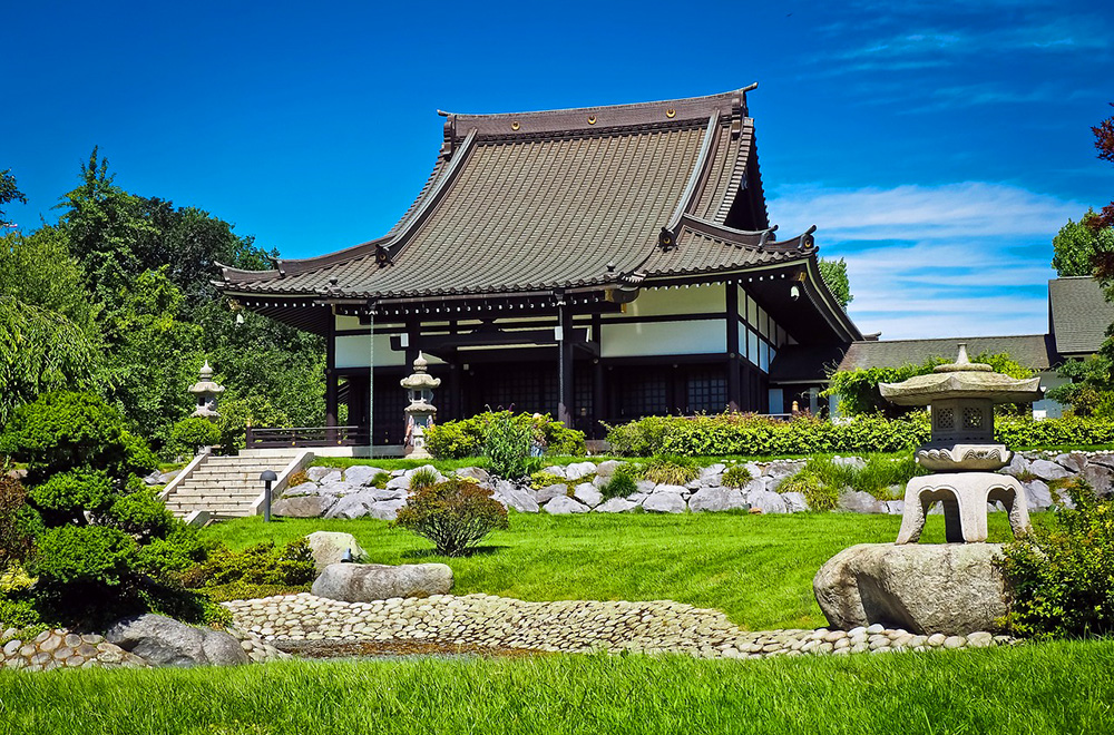A traditional Japanese temple surrounded by a manicured garden with stone lanterns and lush greenery under a clear blue sky.