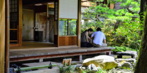 Two people sitting together on the wooden engawa (veranda) of a traditional Japanese house, overlooking a serene garden with lush greenery and stones.