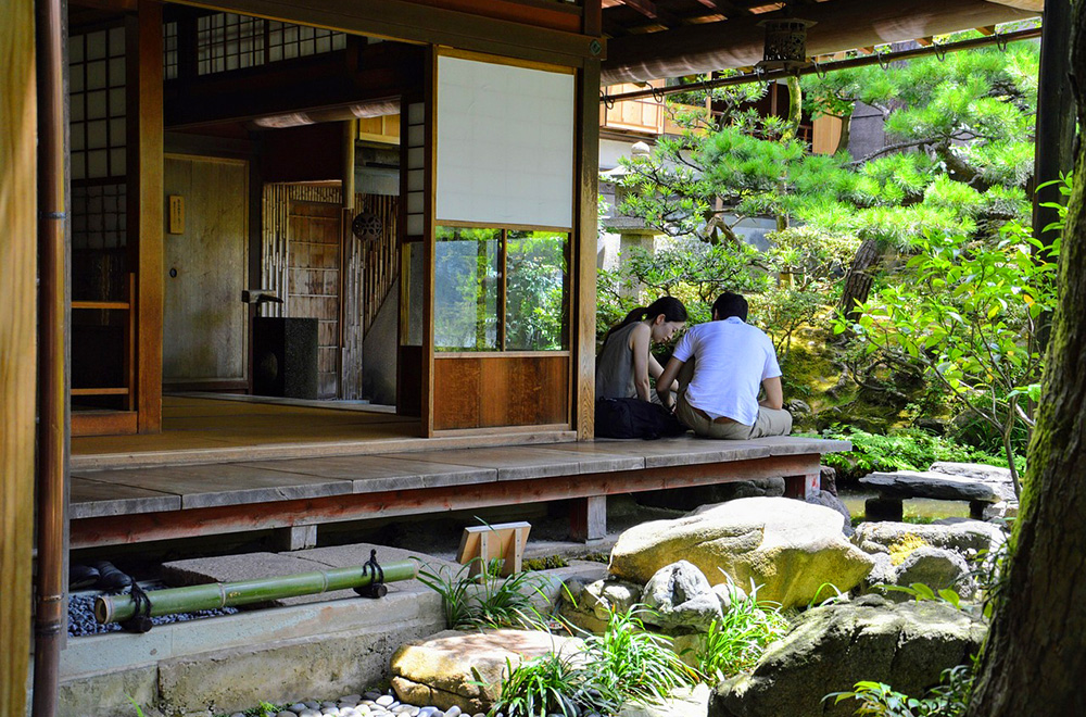 Two people sitting together on the wooden engawa (veranda) of a traditional Japanese house, overlooking a serene garden with lush greenery and stones.
