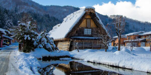 Snow-covered thatched-roof house with a reflection in a pond, surrounded by winter mountains.