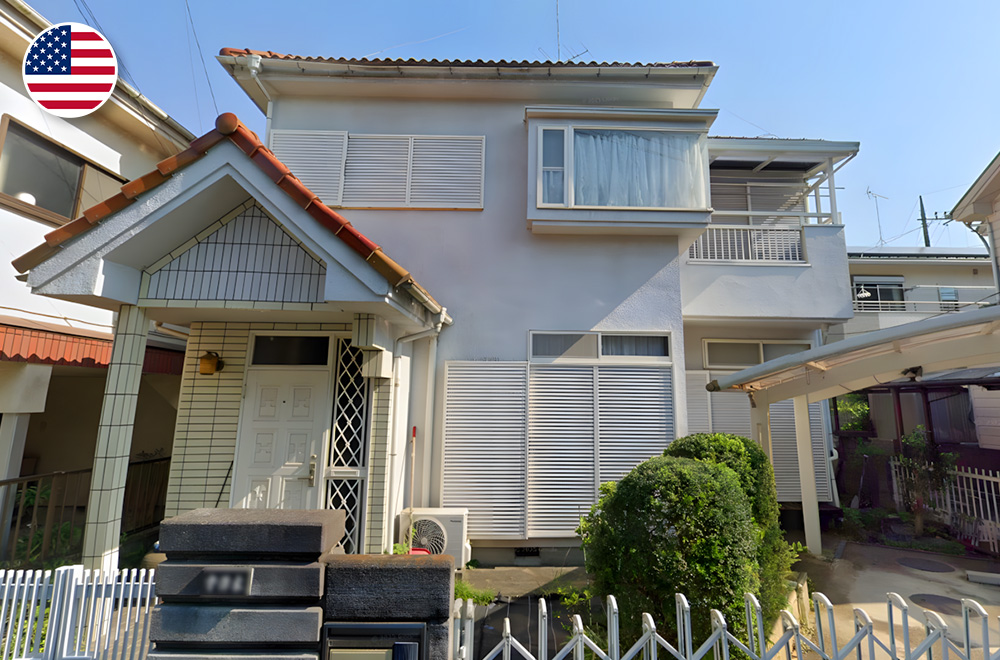 Two-story white house in Japan with tiled entrance roof and closed shutters, small garden, and carport on the right side.