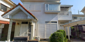 Two-story white house in Japan with tiled entrance roof and closed shutters, small garden, and carport on the right side.