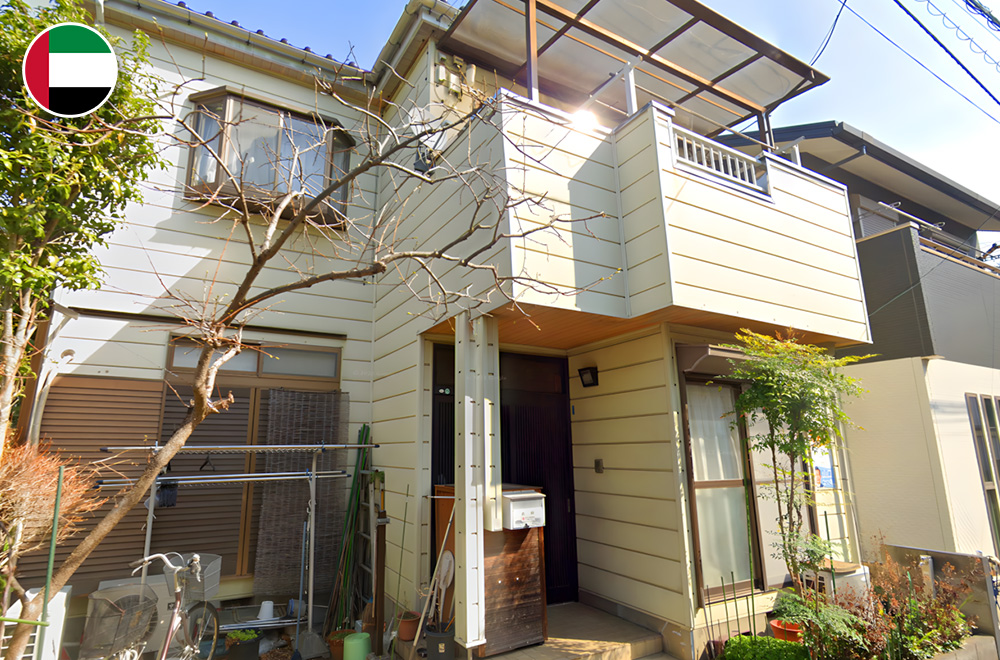 A two-story Japanese house with cream-colored siding, a small balcony, and a simple entrance surrounded by plants and trees on a sunny day.