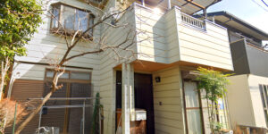 A two-story Japanese house with cream-colored siding, a small balcony, and a simple entrance surrounded by plants and trees on a sunny day.