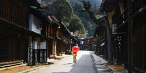 A person in a traditional kimono walks alone down a quiet historic Japanese street, holding a bright red umbrella between dark wooden houses, with forested mountains in the background.