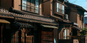 Traditional Japanese residential houses with tiled roofs, wooden facades, balconies, and visible utility wires, illuminated by warm sunlight.