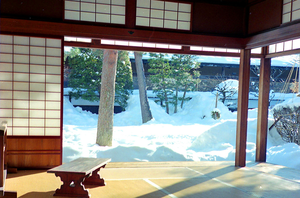 Traditional Japanese tatami room with shoji screens opening onto a snow-covered garden with trees in winter.
