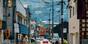 A narrow Japanese town street with small shops, parked cars, overhead power lines, and a pedestrian walking toward the camera, with forested mountains in the background.