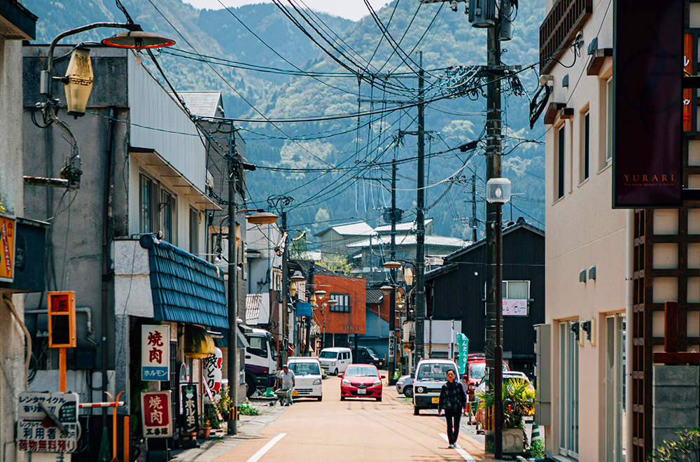 A narrow Japanese town street with small shops, parked cars, overhead power lines, and a pedestrian walking toward the camera, with forested mountains in the background.