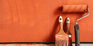 Paint roller and two paintbrushes covered in orange paint leaning against an orange-painted wall on a concrete floor.