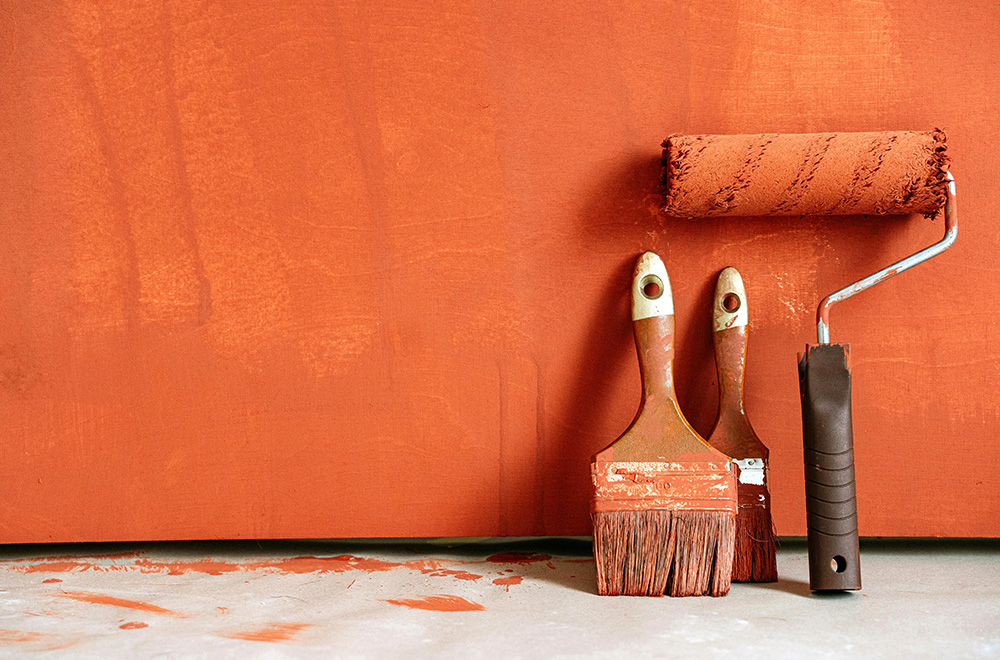 Paint roller and two paintbrushes covered in orange paint leaning against an orange-painted wall on a concrete floor.