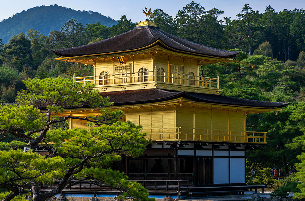 Kinkaku-ji, the Golden Pavilion in Kyoto, a gold-leaf-covered Zen temple surrounded by green trees and forested hills.