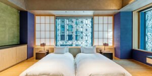 Modern Japanese-style bedroom with two low beds, white bedding, wooden floors, shoji-style panels, and a decorative patterned window.