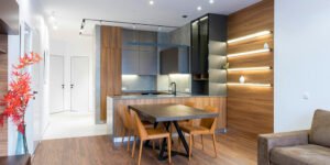Modern open-plan living and dining area with a wooden dining table, four chairs, built-in kitchen, warm wood finishes, and recessed ceiling lighting.