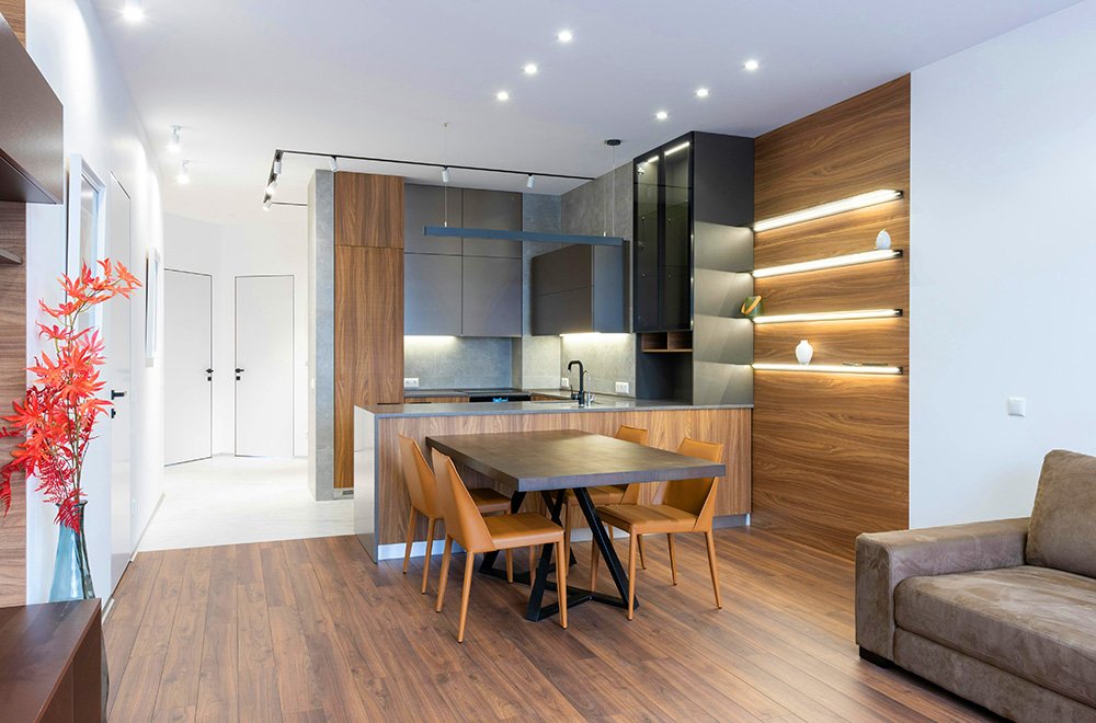 Modern open-plan living and dining area with a wooden dining table, four chairs, built-in kitchen, warm wood finishes, and recessed ceiling lighting.