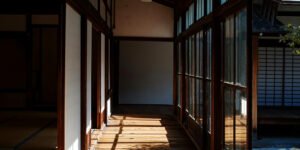 Traditional Japanese wooden corridor with sliding glass doors, tatami room entrance, and sunlight casting long shadows on the floor.