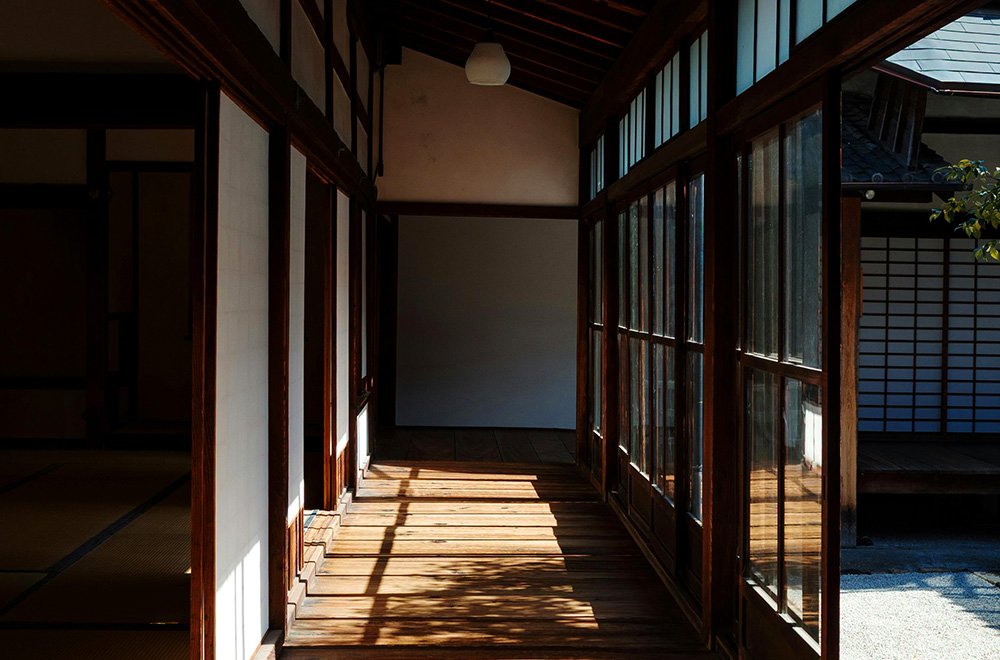 Traditional Japanese wooden corridor with sliding glass doors, tatami room entrance, and sunlight casting long shadows on the floor.