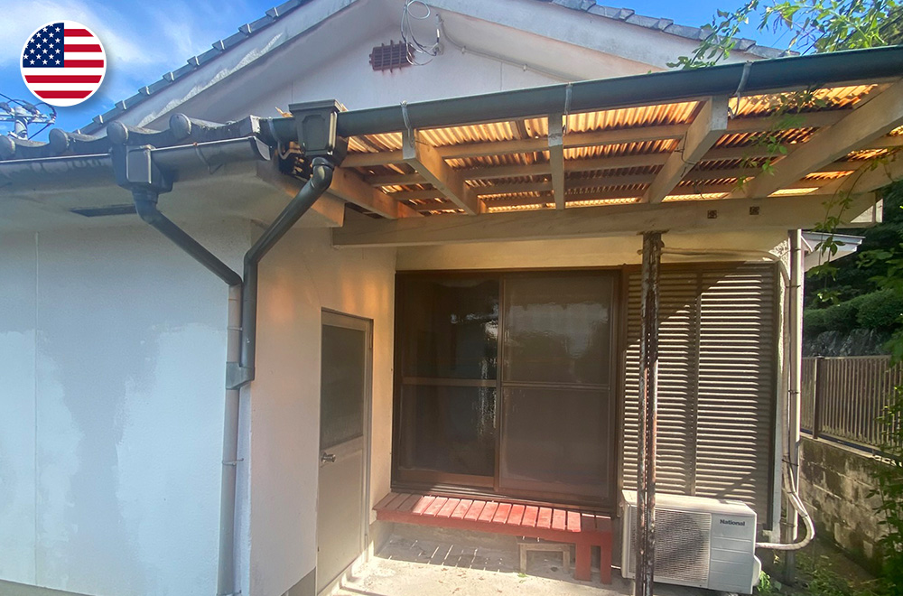 Exterior view of a traditional Japanese house with a tiled roof, wooden-framed sliding window, small awning with translucent panels, and an outdoor air conditioning unit mounted on the wall.
