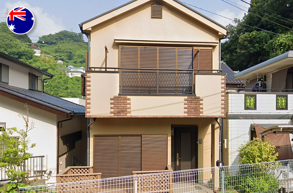 Front view of a two-story Japanese detached house with a beige exterior, brown shutters, a second-floor balcony, and a small fenced front yard, surrounded by neighboring homes and lush green hills.