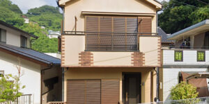 Front view of a two-story Japanese detached house with a beige exterior, brown shutters, a second-floor balcony, and a small fenced front yard, surrounded by neighboring homes and lush green hills.